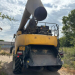 Rear view of a yellow construction excavator with a large black discharge chute, parked on a dirt path among bushes and trees.