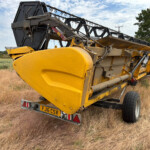 Trailer-mounted yellow combine header in a dry field, with blue sky and green trees in the background, ready for transport.