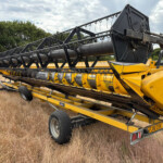 Yellow combine harvester header mounted on a trailer in a dry field with trees in the background, ready for transport.