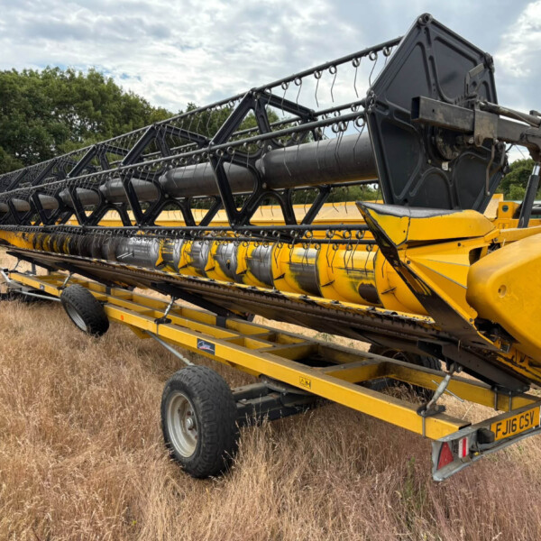 Yellow combine harvester header mounted on a trailer in a dry field with trees in the background, ready for transport.