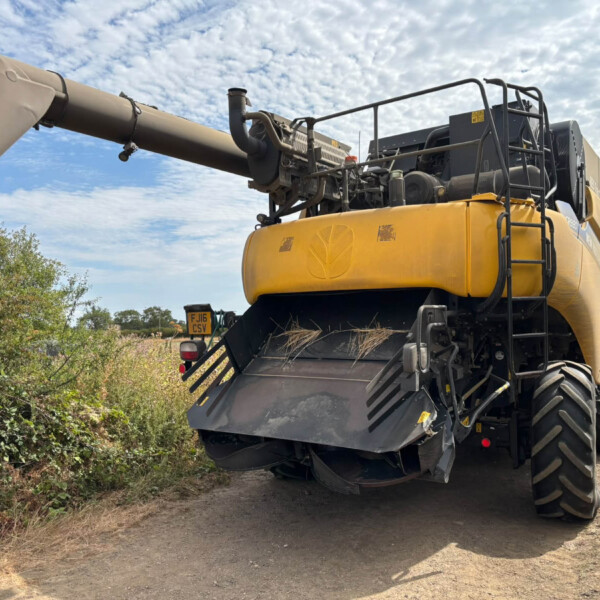 Large yellow construction machine with a long discharge chute extending to the left, on a dirt road with bushes and a blue-sky backdrop.