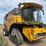 Yellow and black New Holland tracked combine harvester parked on a dirt path in a rural field, with a blue sky above and power lines in the background
