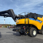 Side view of a yellow New Holland CR9.90 combine harvester with a large black auger extending to the left under a clear blue sky.