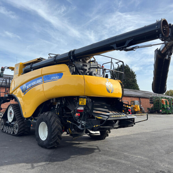 Yellow New Holland CR9.90 combine harvester with a long unloading auger extended into the air in a truck yard.