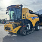 Agricultural combine harvester in yellow, parked beside a building with oversized tires and crop headers attached at the front.