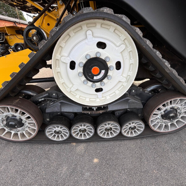 Close-up of a tracked construction vehicle showing a large white road wheel with bolts, belts, and yellow machine frame in the background on pavement.