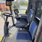 Inside a tractor cab with a steering wheel, blue-and-gray seats, and multiple control panels, monitors, and a joystick for operation.