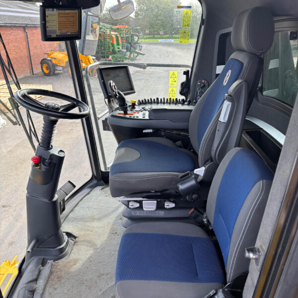 Inside a tractor cab with a steering wheel, blue-and-gray seats, and multiple control panels, monitors, and a joystick for operation.