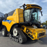 Yellow New Holland combine harvester with tracked wheels and front header parked in an outdoor lot, cab and machinery visible against a blue sky.