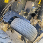 Close-up of a farm trailer undercarriage showing a hitch, large treaded wheel, and debris with yellow warning stickers on a dirty metal frame.