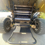 Underside of a large farming header with rollers and belts, hay stuck between components, and big rear tires visible on both sides