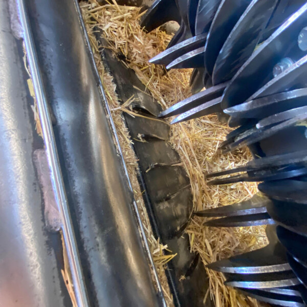 Interior of a metal machine with sharp blades and packed straw or chaff between parts, suggesting a grain or hay processing area.