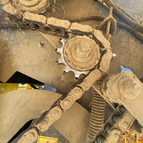 Dusty close-up of a muddy machine undercarriage with a sprocket, chain links, and hydraulic hoses visible under a track system.
