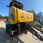 New Holland baler parked in a farmyard under a clear blue sky, with a silo and farm buildings in the background
