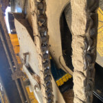 Close-up of a dirty tracked undercarriage (bulldozer/ excavator) with metal rollers and drive chains, covered in mud and straw debris.