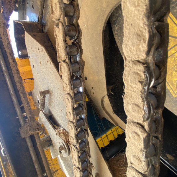 Close-up of a dirty tracked undercarriage (bulldozer/ excavator) with metal rollers and drive chains, covered in mud and straw debris.