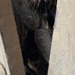 Close-up of a dusty, dark wooden wheel or cog lodged between two weathered wooden beams with straw debris tangled in the gap.