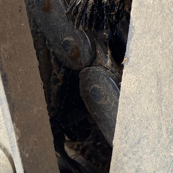 Close-up of a dusty, dark wooden wheel or cog lodged between two weathered wooden beams with straw debris tangled in the gap.