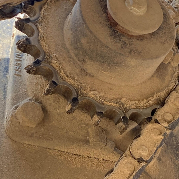 Close-up of a dusty, heavy-machinery gear or sprocket with jagged teeth and a circular hub, partially buried in soil during excavation.