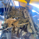 Close-up of a dusty harvest machine's gears, belts, and hydraulic parts with straw wedged in the mechanism.