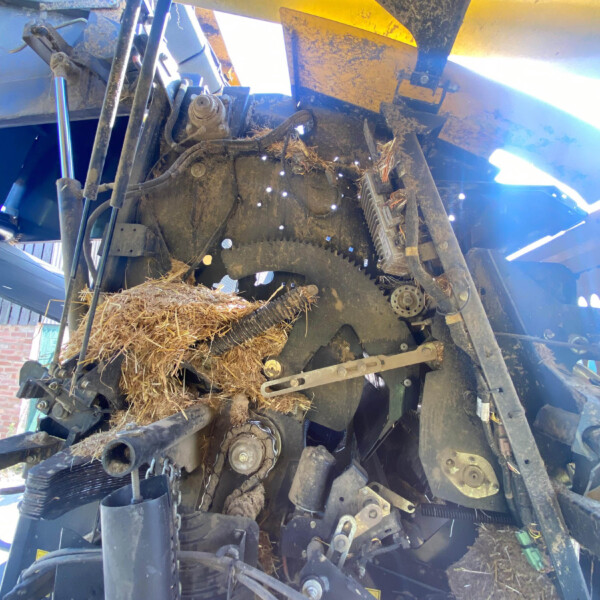 Close-up of a dusty harvest machine's gears, belts, and hydraulic parts with straw wedged in the mechanism.