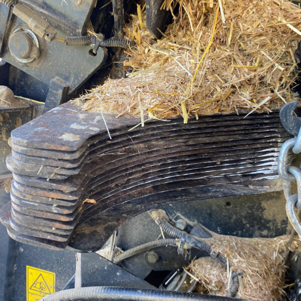 Close-up of a hay baler with straw packed around gears and hydraulic hoses, safety sticker visible near the bottom left.