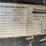 Close-up of a metal identification plate on agricultural equipment showing CNH Industrial Belgium NV and New Holland Agriculture branding with product and load specs visible in dirt.