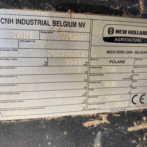 Close-up of a metal identification plate on agricultural equipment showing CNH Industrial Belgium NV and New Holland Agriculture branding with product and load specs visible in dirt.