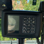 Operator control panel with numerous labeled buttons and a small screen inside a vehicle cab, with a tethered cable visible outside the window.
