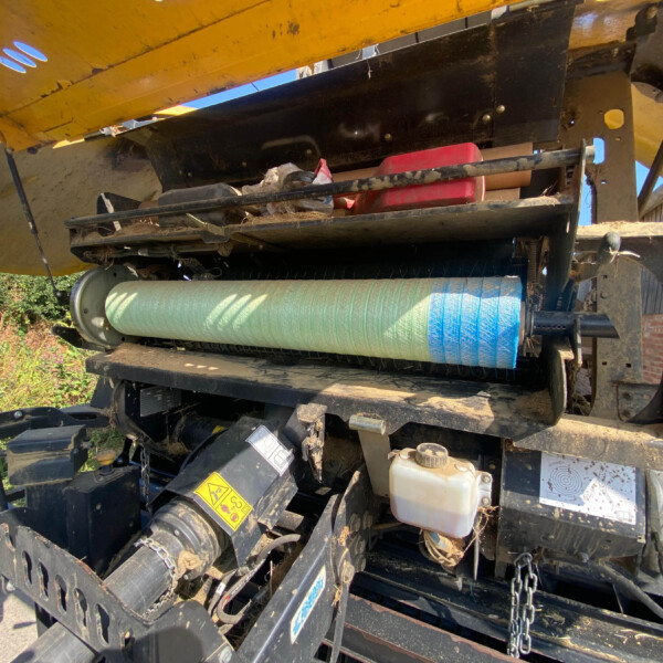 Close-up view of a yellow agricultural baler showing belts, a dusty black frame, a green horizontal roller, and a red item resting inside the open housing, outdoors.