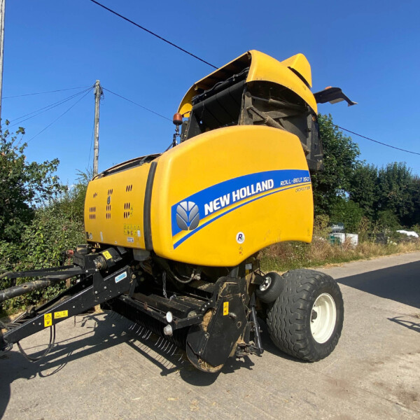 New Holland Roll-Belt 150 forage harvester with the front header raised, parked on a road against a clear blue sky.
