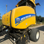 Yellow New Holland Roll-Belt 150 agricultural harvester viewed from the side, logo and blue stripe visible on the body behind the cab window area.