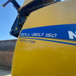 Close-up of a yellow agricultural harvester with blue stripe reading Roll-Belt 150 and Crop Cutter, against a clear blue sky.