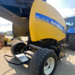 Front view of a yellow New Holland Roll-Belt 150 Crop Cutter header attached to a blue harvester, with large tires visible and a blue stripe label.