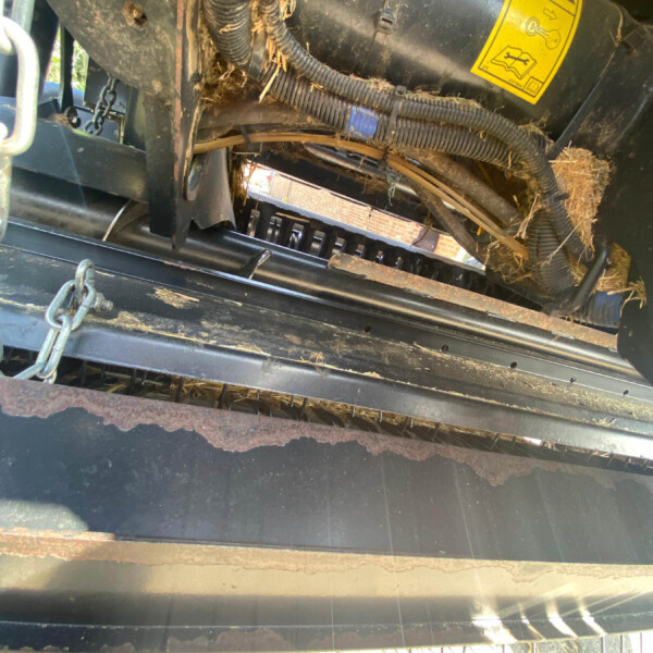Close-up of a dusty agricultural mower mechanism with bundled wires, straw, and a yellow safety label.