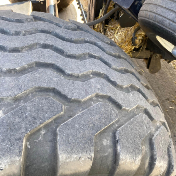 Close-up of a large tractor/trailer tire with deep, zigzag tread, with hay and undercarriage components visible behind it on a farm trailer.