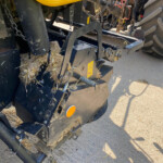 Close-up of a yellow farm implement with black metal frame and straw caught in the machinery, showing chains and warning decals.