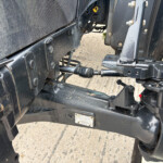 Close-up of a tractor's rear hitch and hydraulic linkage, with a large tire visible on the right and dusty metal parts nearby.