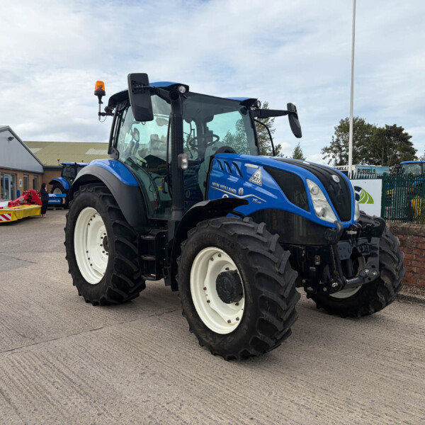 Blue New Holland tractor photographed from the front side, parked outside a building with safety barriers nearby and a blue flag in the background.