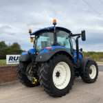 Front-left view of a blue agricultural tractor with orange beacon lights, parked near a brick wall and power lines under a cloudy sky.