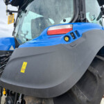 Close-up of the blue tractor front with a large black hood, partially visible wheel, and warning stickers on the body under the windshield.