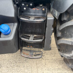 Close-up of a dirty tractor footstep with a rusted, worn tread and nearby blue coolant cap on the left.