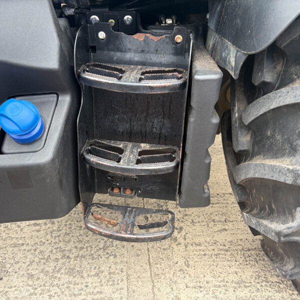 Close-up of a dirty tractor footstep with a rusted, worn tread and nearby blue coolant cap on the left.