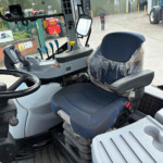Cab interior of a blue tractor showing the driver’s seat with plastic cover, steering wheel, and colorful levers, view through the windshield to a yard with tractors outside.