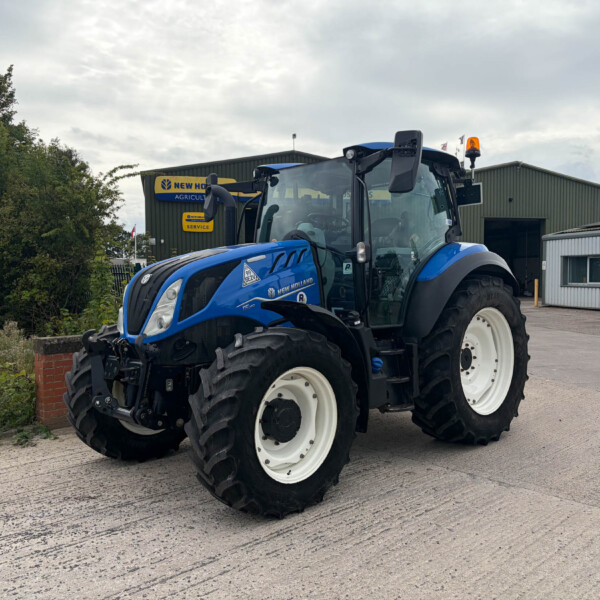 Blue New Holland tractor parked outside an agricultural service building with a cloudy sky above.