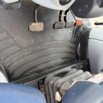 Close-up of three metal motorcycle foot pedals with diamond-pattern grips, mounted above a ribbed rubber floorboard in a dusty scooter interior.