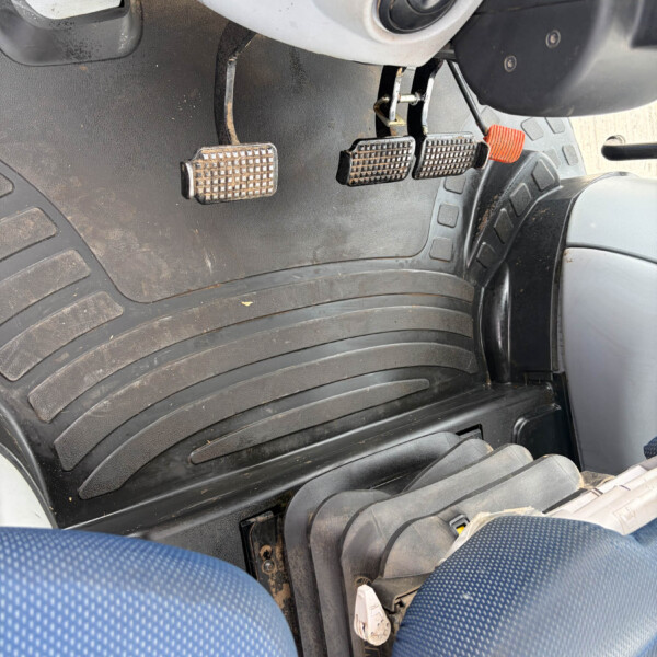 Close-up of three metal motorcycle foot pedals with diamond-pattern grips, mounted above a ribbed rubber floorboard in a dusty scooter interior.