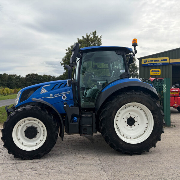 Close-up of a blue New Holland tractor at a dealership yard, showing large white wheels and front tire treads.