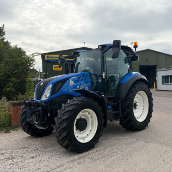 Blue New Holland tractor in foreground with a second tractor and a warehouse in the background.