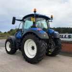 Front-left view of a blue agricultural tractor with large white wheels and orange beacon lights in a parking yard outdoors on a cloudy day